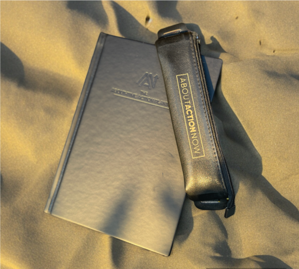 Black notebook titled "The About Action Planner" partially covered by a black pencil case with the text "ABOUT ACTION NOW" lying on a sandy surface.