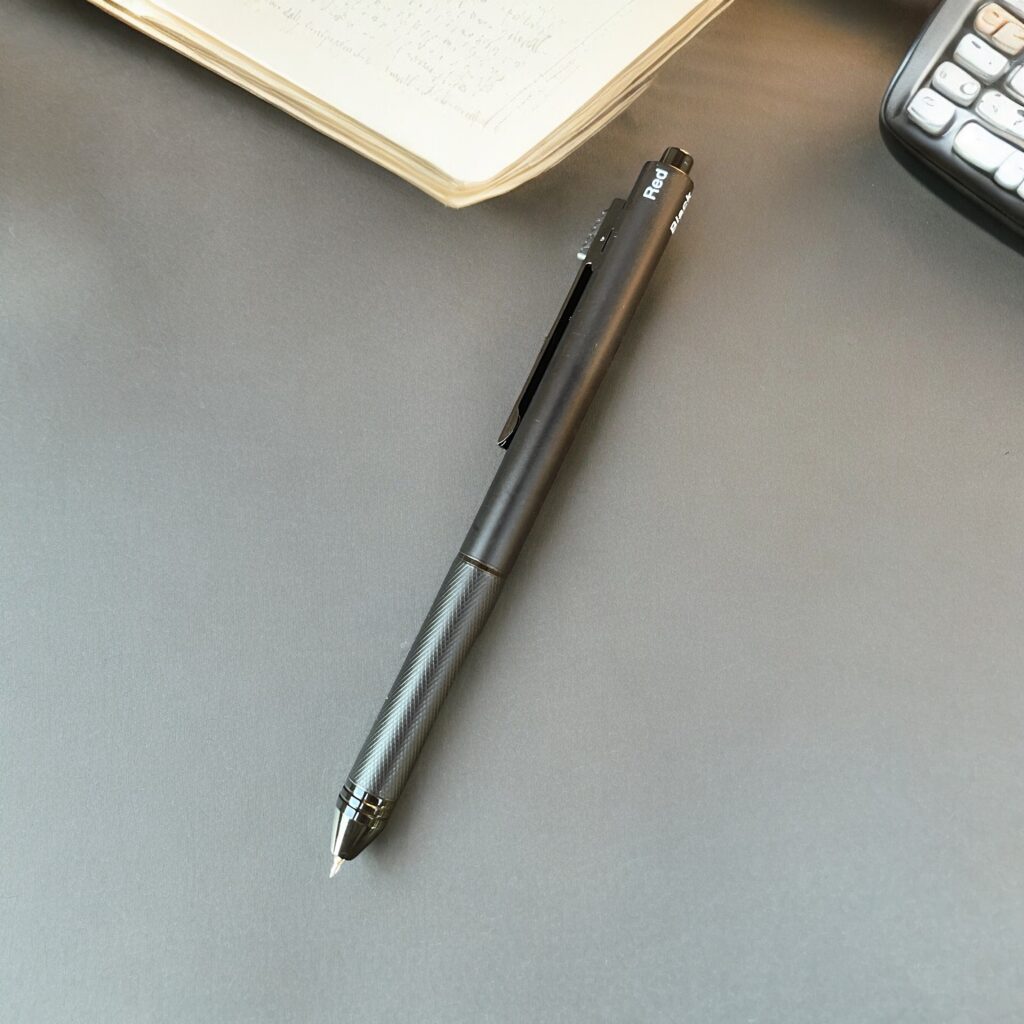 Black About Action Now multi-functional pen lying on a grey desk with an open notebook and a calculator in the background.